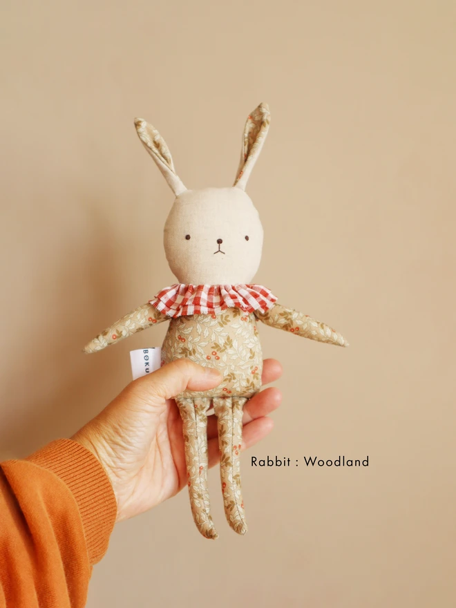 Handmade rabbit doll with light floral-patterned fabric, a red-and-white gingham ruffle collar. A white tag with the brand name ‘BOKUNO’ is sewn on the side.