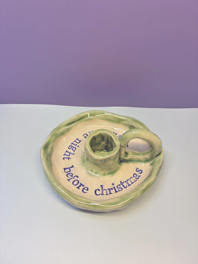 Handmade ceramic Christmas taper candlestick holder with a soft green glazed edge and the words “‘Twas the night before Christmas” in dark blue lettering, arranged around the candle holder opening. Photographed against a pale background.