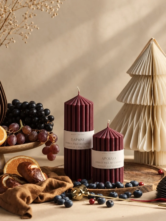 Daphne & Apollo Burgundy Pair Candles (Set Of 2). The tall Daphne and shorter Apollo ribbed pillar candles in deep burgundy, on a wooden tray with blueberries, cranberries, a brass snuffer and a velvet ribbon in warm sunlight. The handmade soy wax candles make an elegant gift for the holiday season.
