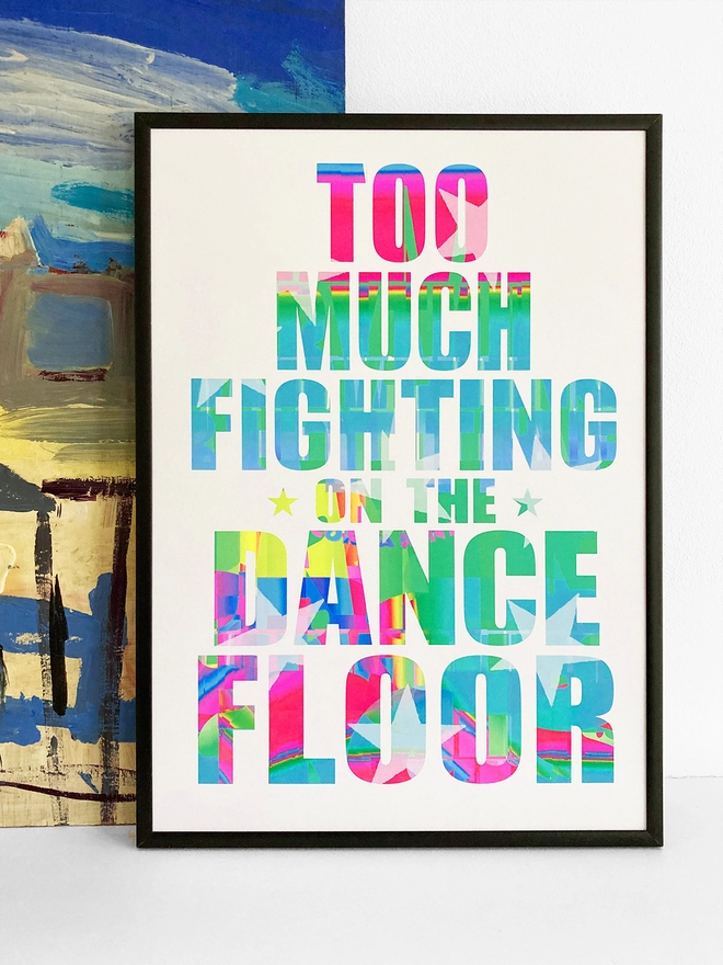 Framed multicoloured typographic print - Lyrics from The Specials song Ghost Town "too much fighting on the dance floor" The print rests against a blue and yellow abstract painting. 