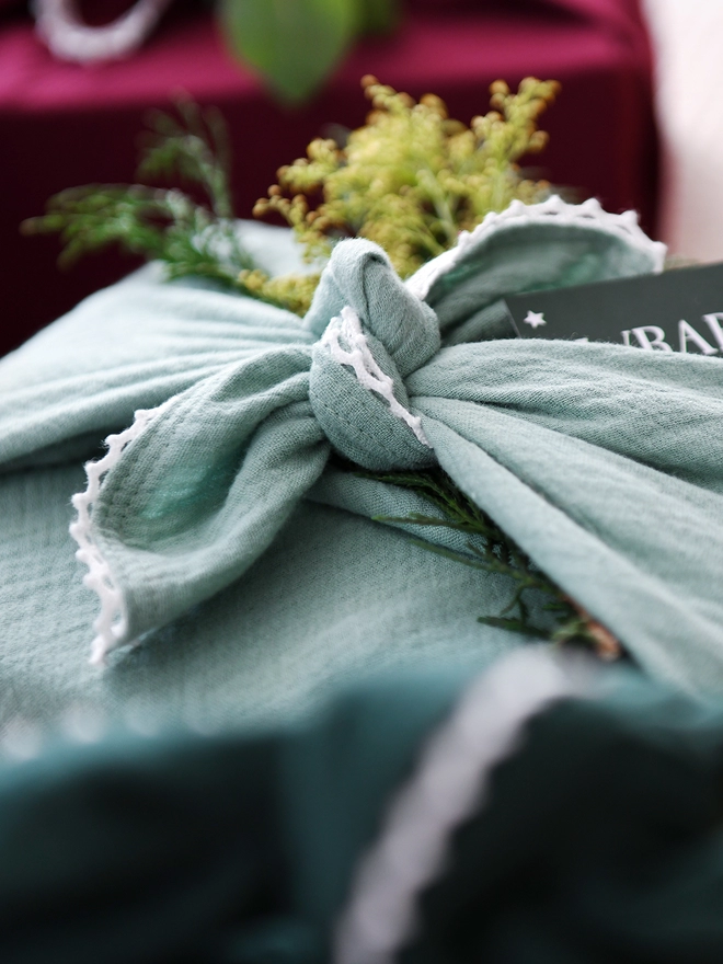 A gift wrapped in mint green cotton fabric wrap with an ivory lace trim, that is knotted on top, has a stem of flowers tucked into the knot.