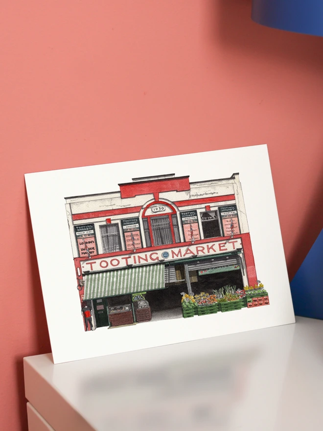 An architectural illustration of the Tooting Market storefront on a print, displayed on a white surface against a pink wall.