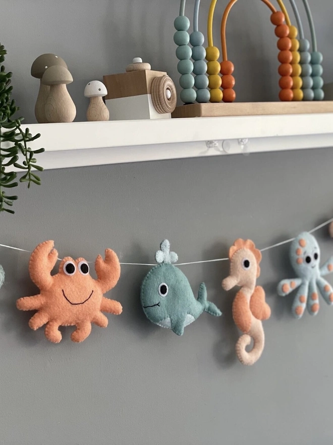 Nursery Under The Sea Garland, a sea themed garland hanging from a white shelf against a grey backdrop in a child's bedroom. 