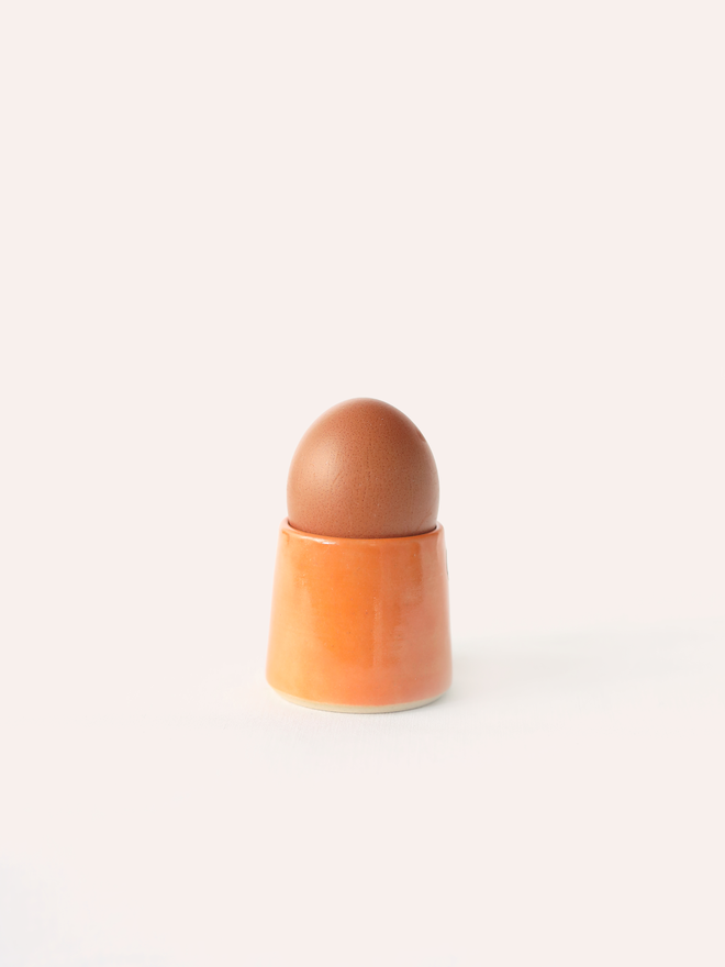 A handmade ceramic egg cup in mandarin orange glaze holding a brown egg, photographed against a plain white background