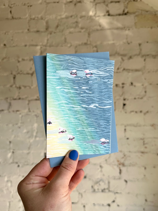 a greetings card featuring an illustration of flamingos in water with a rainbow reflection with a blue envelope