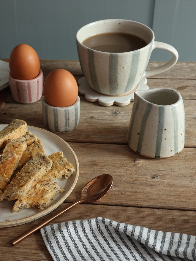 A table scene showing two handmade ceramic egg cups with eggs – one with pink stripes and one with blue and white stripe - a mug and milk jug in blue and white stripe, and a plate with toast. A brass spoon and blue and white striped napkin can be seen in the foreground. 