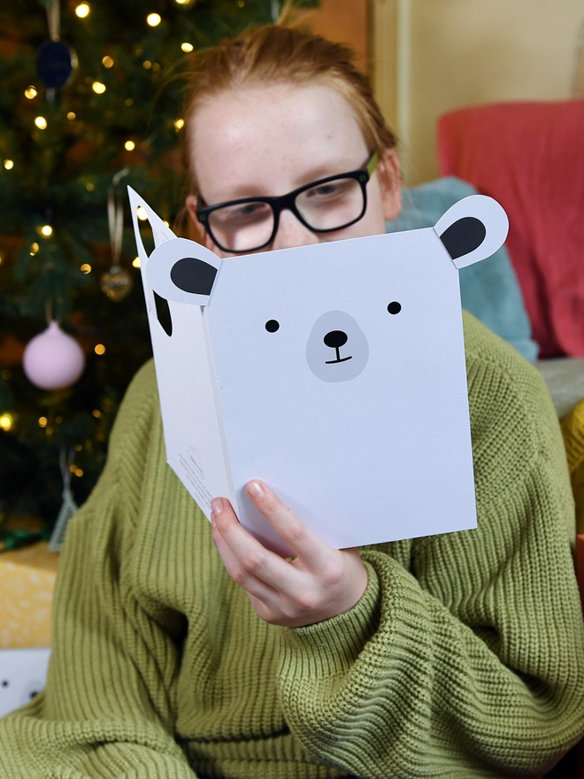 A child wearing a green jumper sits in front of a Christmas tree and holds a polar bear card in front of them.