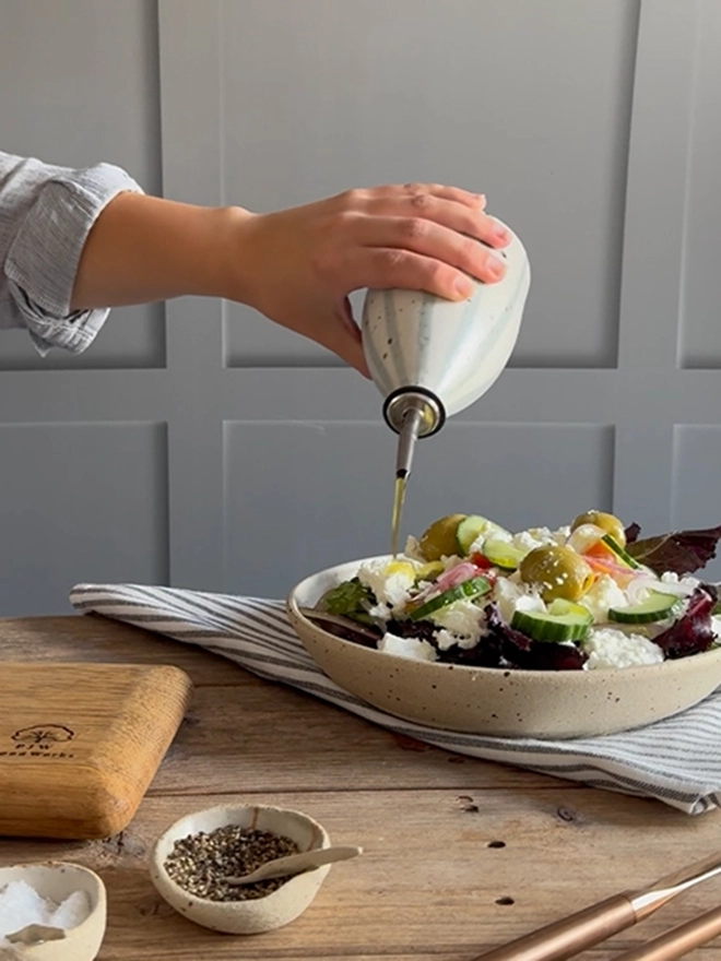 A female hand holding our Handmade Ceramic Stoneware Oil Pourer in Blue and White Stripe and pouring oil from the bottle over a salad. The bowl of salad sits on a blue and white striped napkin on a wooden surface, alongside small ceramic dishes containing salt and pepper, brass cutlery and a wooden chopping board. 