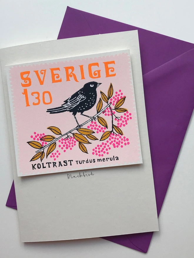 Bird Stamp Card Swedish/ Blackbird, a white card with a colourful bird design on a white backdrop. It sits above a purple envelope.