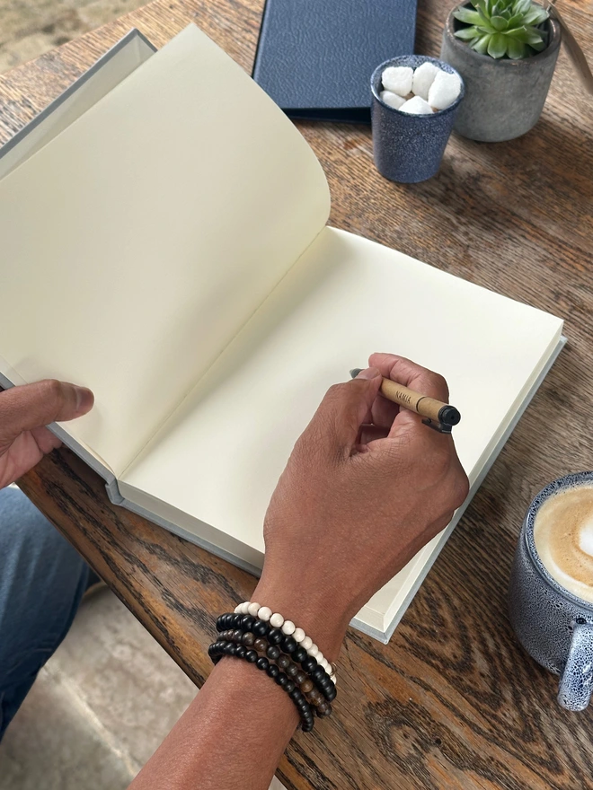 a man is holding a pen in his hand and he is about to write in a notebook. The notebook has plain paper. In the background is a cup of coffee.