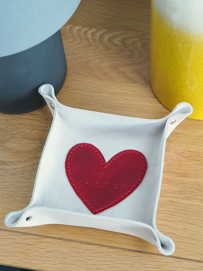 Personalised leather heart coin tray. A natural cream square leather tray with a red leather heart stitched to the base. The heart is personalised with a couples initials and their wedding date.