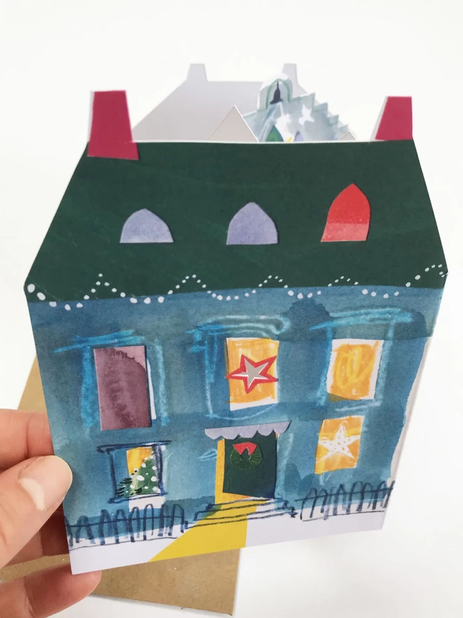 Hand holds illustrated cut-out card showing festive house with lit-up windows