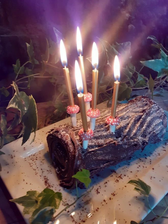 Handmade ceramic fungi-inspired cake decorations illuminating a chocolate yule log with five slender gold candles, framed by trailing green ivy.