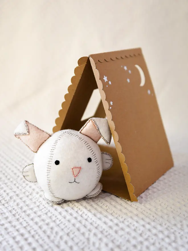 A white felt rabbit toy sits on a fabric surface beside a cardboard house.