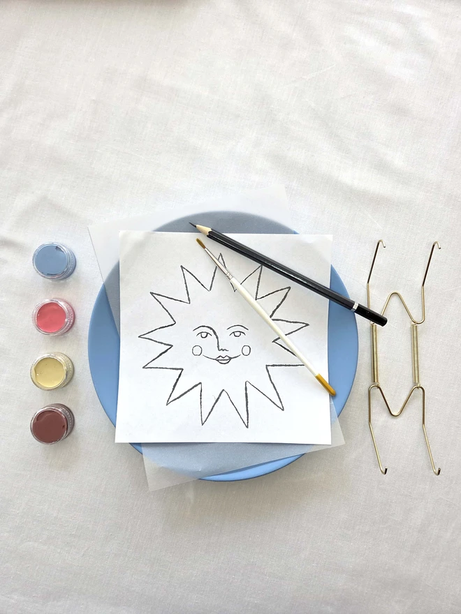Contents of the sun plate painting kit on a white cloth - plate, design template, brush, pencil, paints and wall hook. 