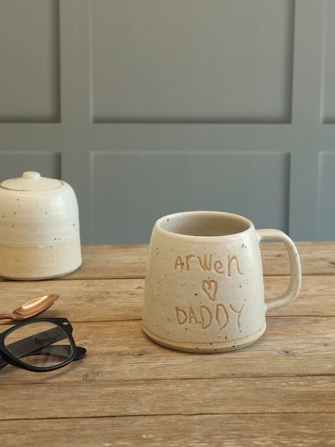 Personalised Handwriting Stoneware mug with the text 'Arwen loves Daddy' in a child's handwriting on the front. The mug is sat on a wooden surface with a matching sugar pot, rose gold spoon and a pair of glasses. 