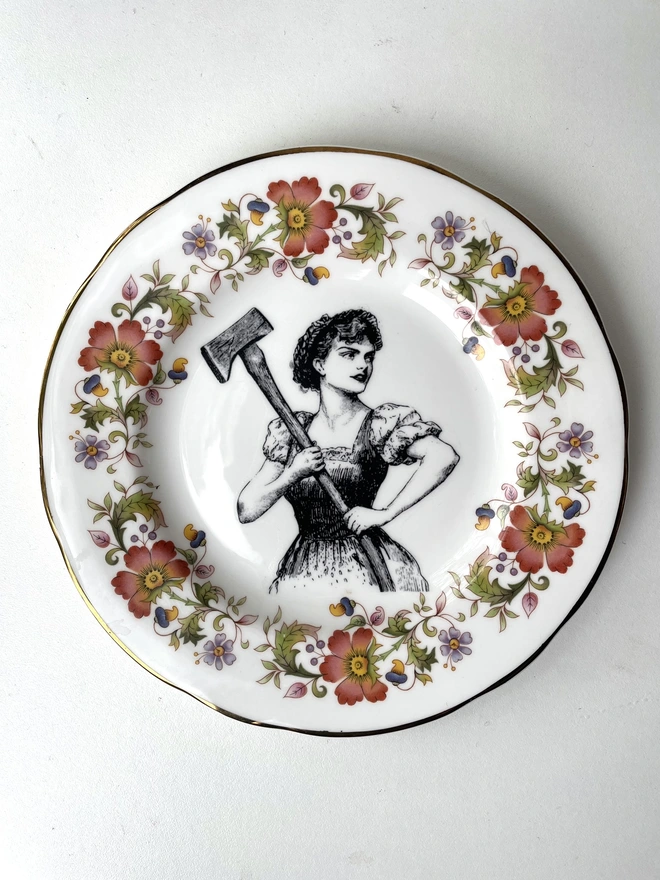 a vintage plate with a really ornate pretty border has a vintage black and white illustration of a beautiful girl in peasant clothes wielding an axe