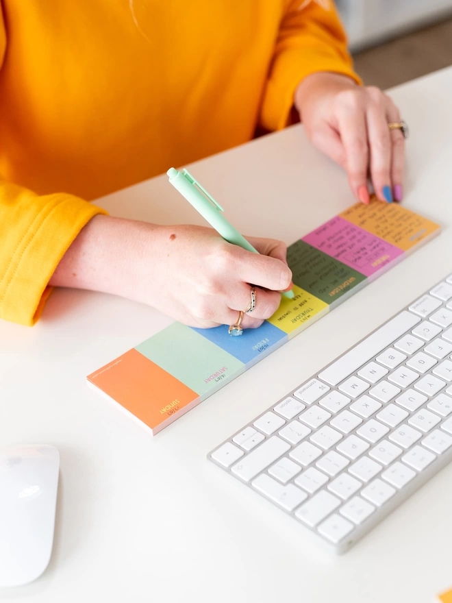 A woman in an orange jumper and colourful rainbow nails fills out the Raspberry Blossom Keyboard Jot Pad
