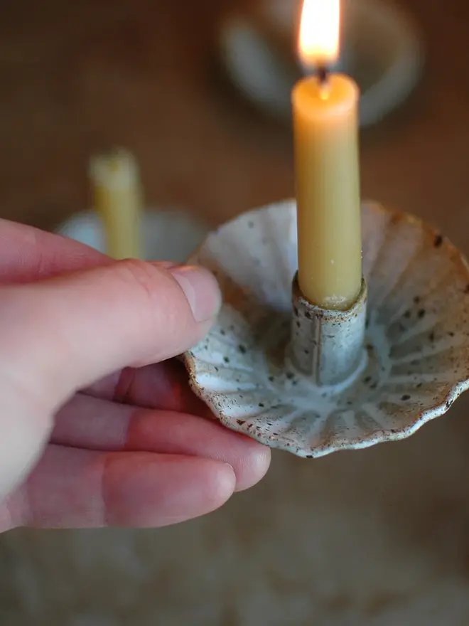 Handmade Natural Speckled Ceramic Candlestick Holder, a person holding a ceramic candlestick holder in the air. It is accompanied by a lit candle. 