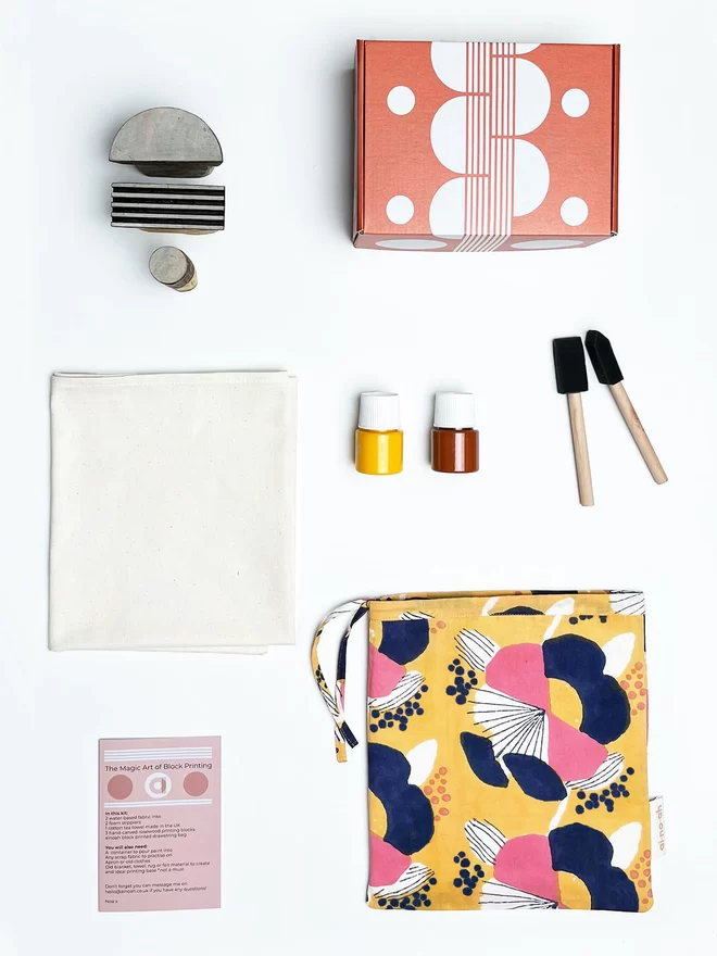 Block printing kit with a printed yellow drawstring bag, two paint pots, one yellow and one cinnamon coloured, two foam brushes, an instruction booklet, three geometric wooden blocks and a plain cream tea towel, next to a rectangular orange box with a white geometric design printed on it on a white surface