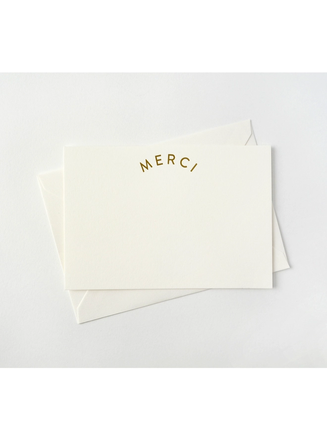 Notecard on white paper that says 'Merci' in Gold Foil, with an Envelope that has a golden seal