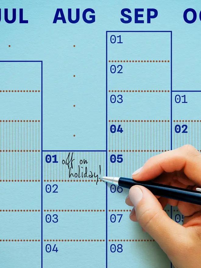 Closeup of a blue 'sky' Dotline 2027 yearly wall planner showing discrete typographic details, a note saying 'off on holiday' and a pen in hand