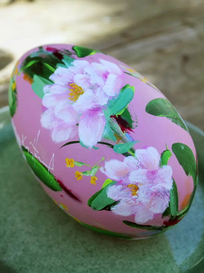 pale pink floral hand painted wooden egg, adorned in pink peonies and yellow cowslip flowers, positioned on a green plate