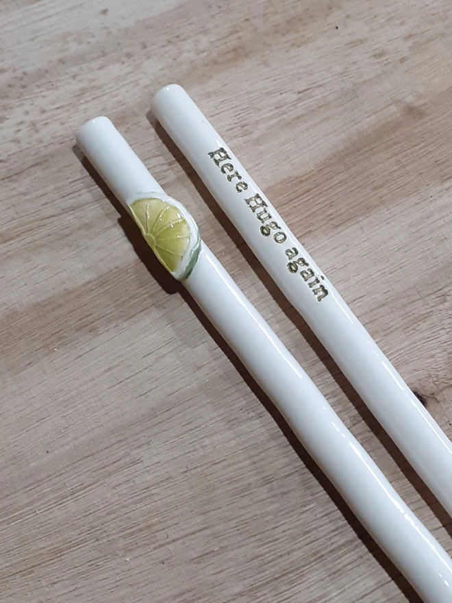 Photo of two handmade ceramic drinking straws on a wooden surface — one decorated with a sculpted lime slice and the other reading “Here Hugo again” in raised lettering, inspired by the Hugo Spritz cocktail.