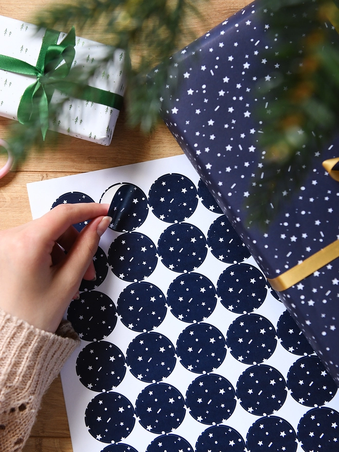 A hand is peeling a navy blue sticker with a tiny star pattern from a sheet of 35 matching stickers.