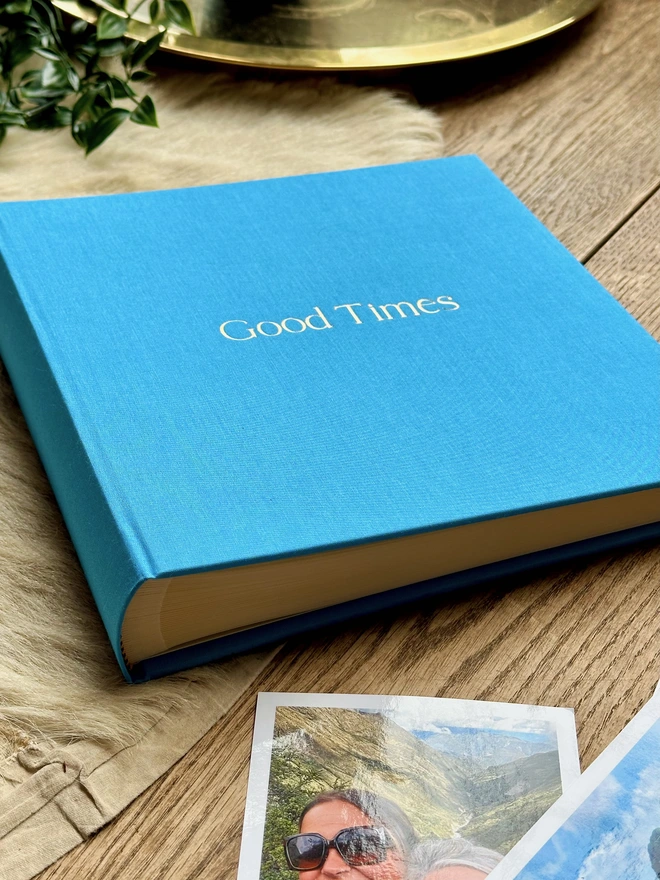 a blue photo album has been printed with the words Good Times on the front. There are lots of photos of friends in the background.
