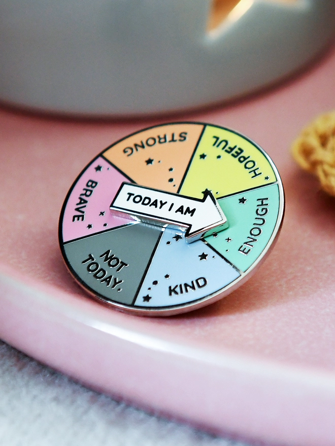 A round enamel pin that looks like a pie chart with six segments, each with positive word, and a white arrow that reads "Today I Am", is resting on a pink plate beside a candle.