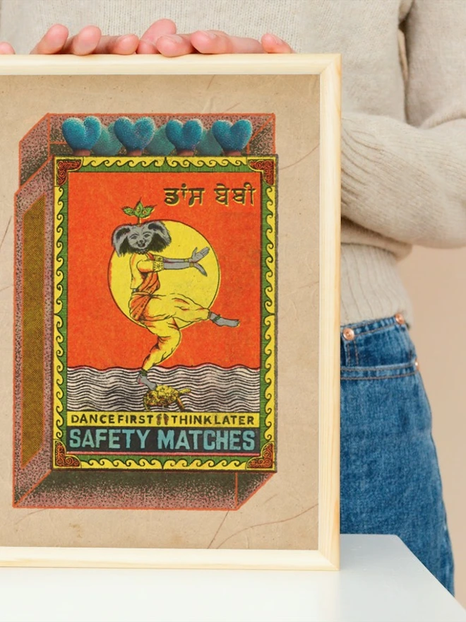 A vintage-style illustrated matchbox design featuring a dancing figure with a dog’s head and a human body dressed in yellow and orange traditional clothing. The figure balances on one leg over water, with a turtle beneath their foot, and a large yellow sun in the background. Above, there are five blue heart-shaped match heads. The matchbox border is decorated with ornate patterns. Text at the bottom reads: “DANCE FIRST THINK LATER SAFETY MATCHES.”