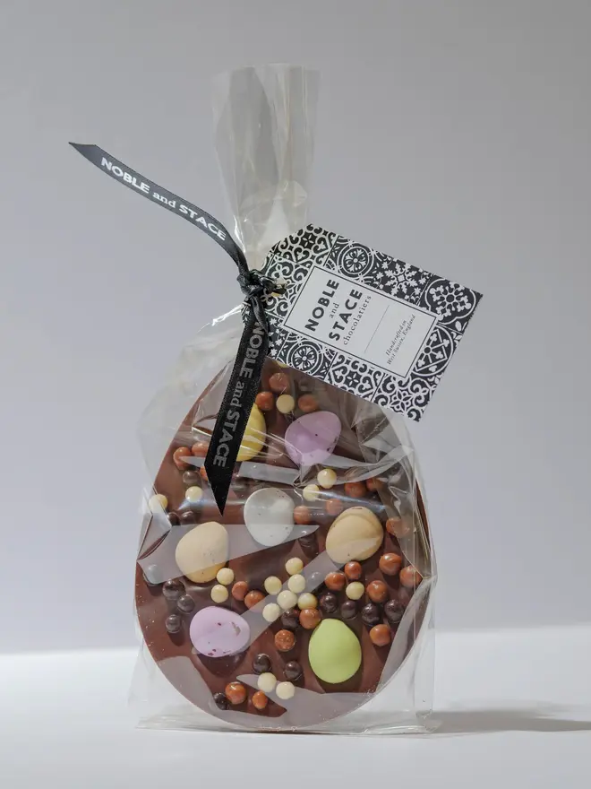 Large milk chocolate egg-shaped tablet decorated with pastel mini eggs and assorted chocolate pearls. Wrapped in clear plastic and standing upright on a plain white background.