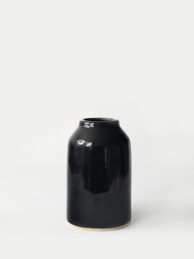 A small handmade ceramic bud vase in glossy jet black glaze, with a rounded bottle shape and narrow neck, photographed against a plain white background