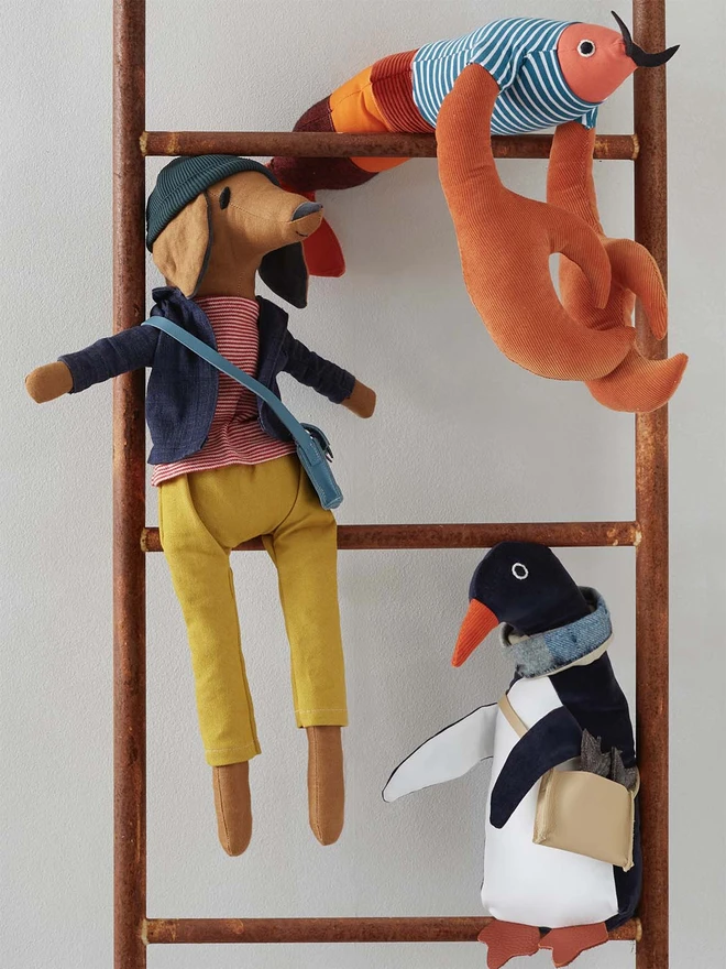 Handmade toy dark blue velvet penguin wearing a tweed and linen scarf and a red leather satchel with three tweed fish in it. On a ladder with a toy lobster and toy sausage dog