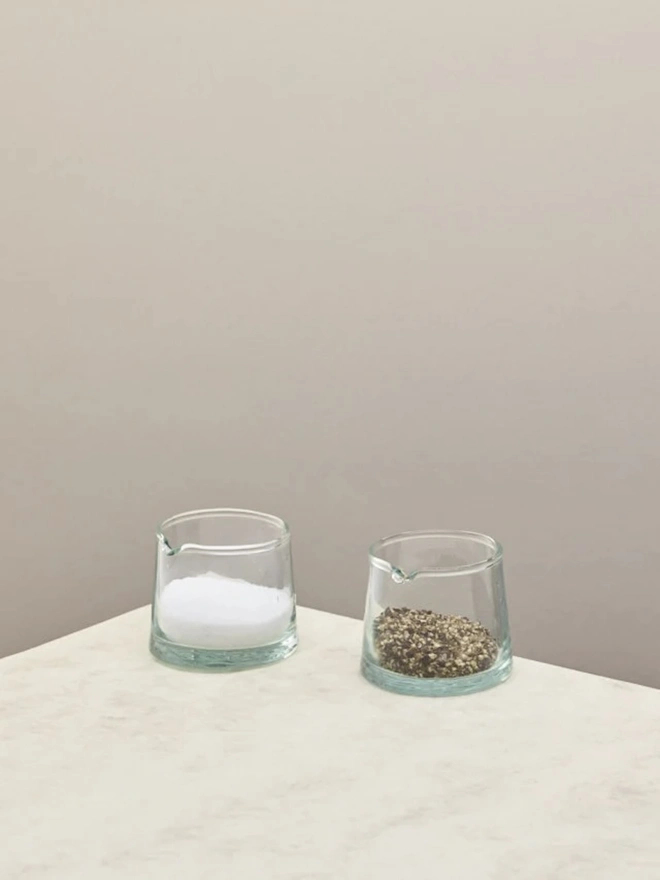 Low Jug, a small glass jug sitting on a counter top against a plain backdrop. It is filled by pepper and accompanied by another jug of salt. 