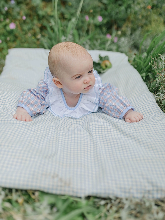 A baby lies on a matress on the ground in a checked romper with a white frilled collar.