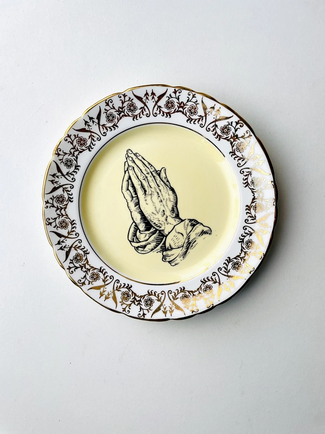 a vintage plate with an ornate white and gold frame, has a yellow centre, and within the centre is a vintage illustration of a pair of praying hands