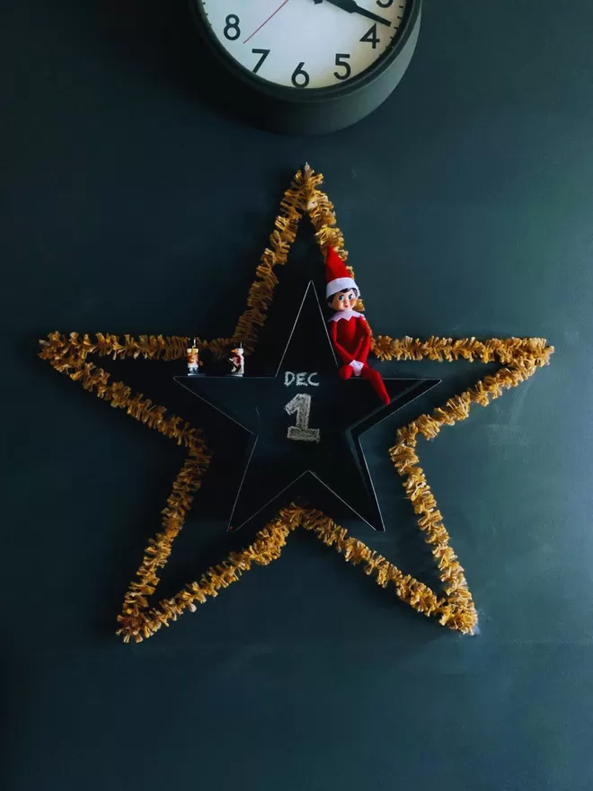 Warm Gold string tinsel AKA Strinsel shaped to form a star on a dark grey wall, with an Elf of the Shelf and Dec 1st written in chalk 