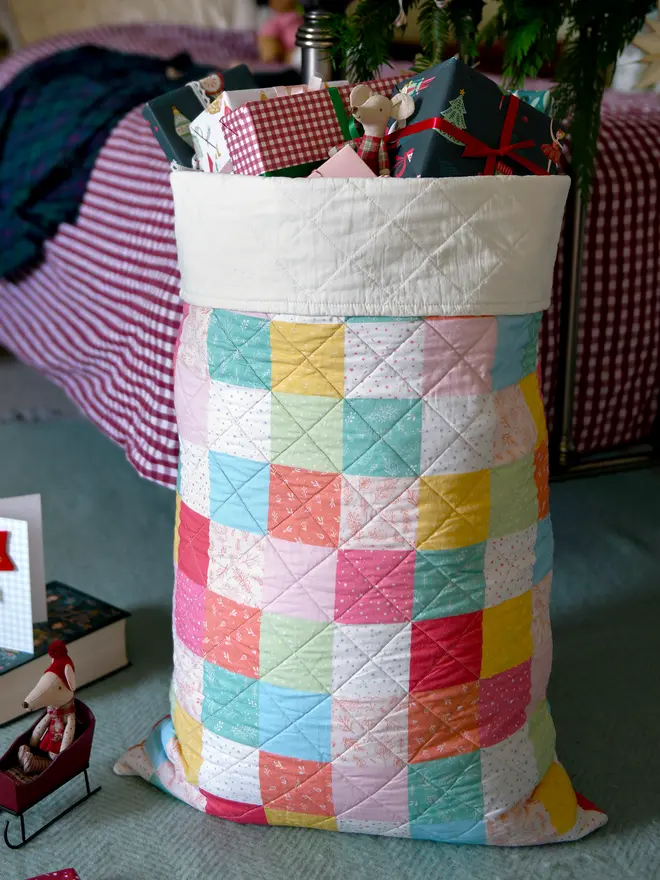 A handmade pastel patchwork Christmas sack stands beside a bed.