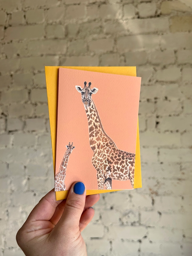 an orange greetings card featuring illustrations of two giraffes with a deep yellow envelope