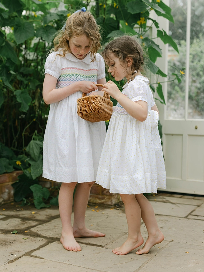 Two girls stand in a green house wearing white dresses with rainbow smocking