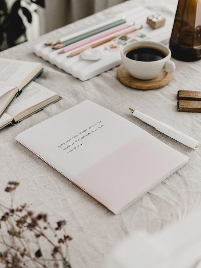 pale pink and white handmade eco-friendly, personalised notebook with text typed on the cover, on a desk with a fountain pen and coffee