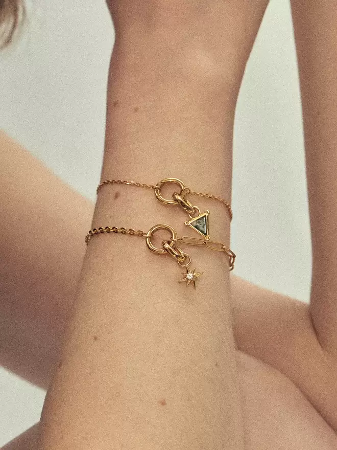 Woman wearing two gold bracelets stayed with a dendritic chaceldony shield charm and a gold star charm