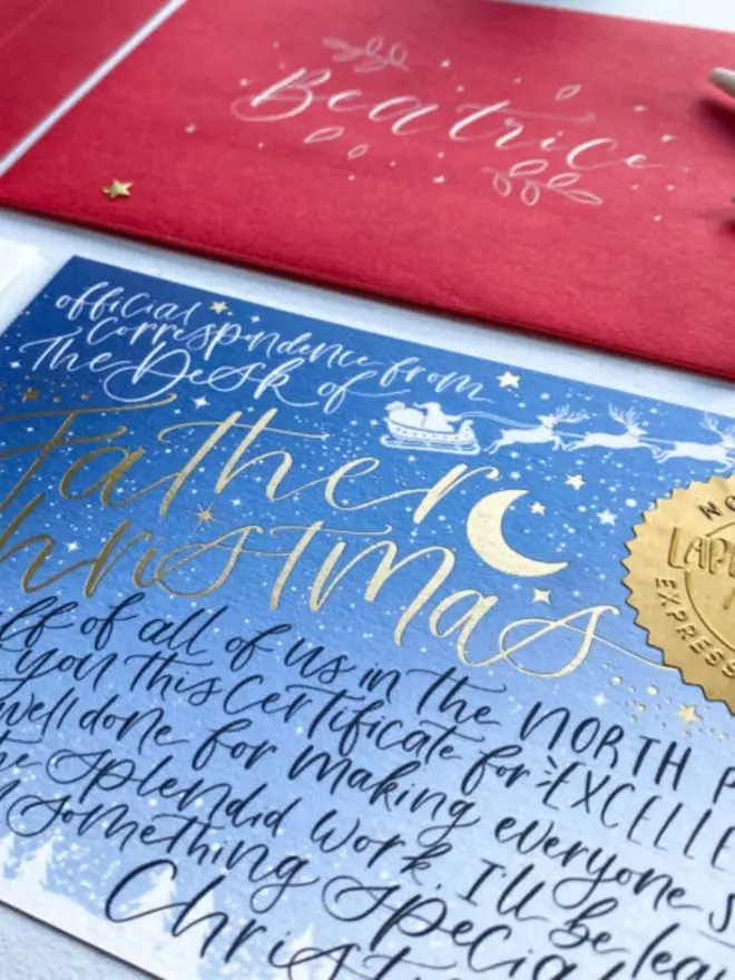 Gold foil "Father Christmas" lettering in front of a red envelope with "Beatrice" hand-lettered in white.