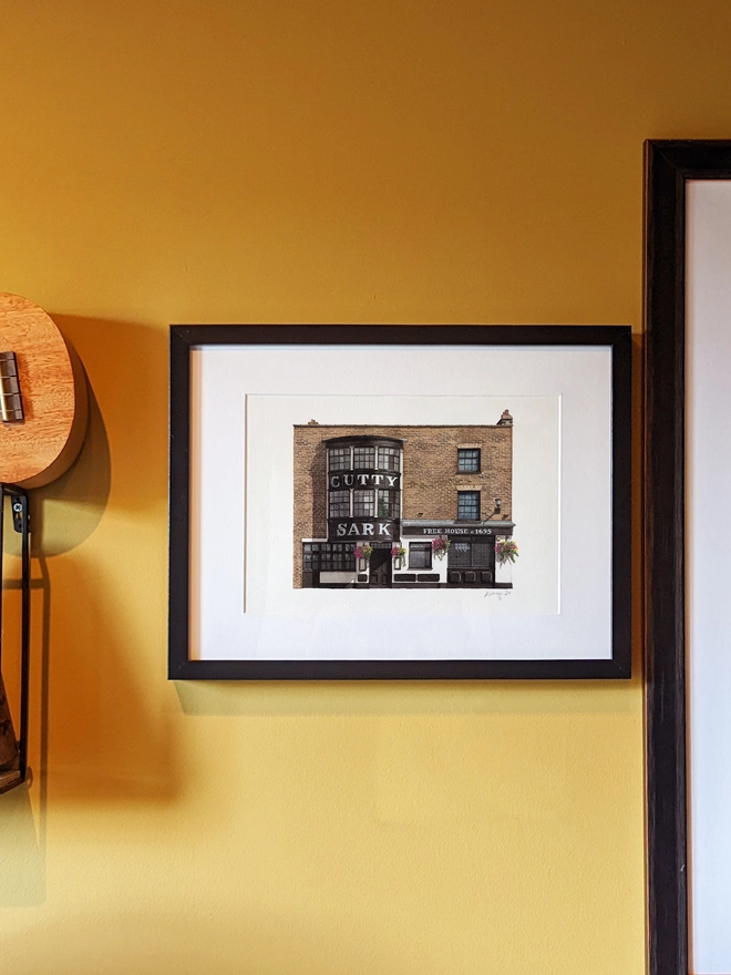 A framed watercolor print of the Cutty Sark pub hanging on a yellow wall next to a ukulele and another framed artwork.