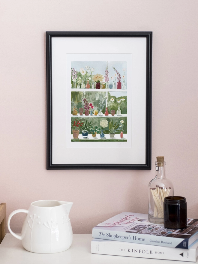 A photograph of an art print on a pale pink wall. The print features shelving within a glasshouse. The white shelves are covered in small glass vessels holding single stems of daisies, foxgloves and hydrangeas. There are also terracotta posts that hold succulents and house plants.