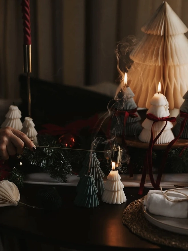 Forest Green Christmas Tree Warm Candles (Set Of 2). A hand snuffing out a green tree candle with a brass snuffer as smoke curls up, beside cream and grey tree candles and a paper honeycomb tree. The scene captures the cosy ritual of candlelit Christmas evenings.