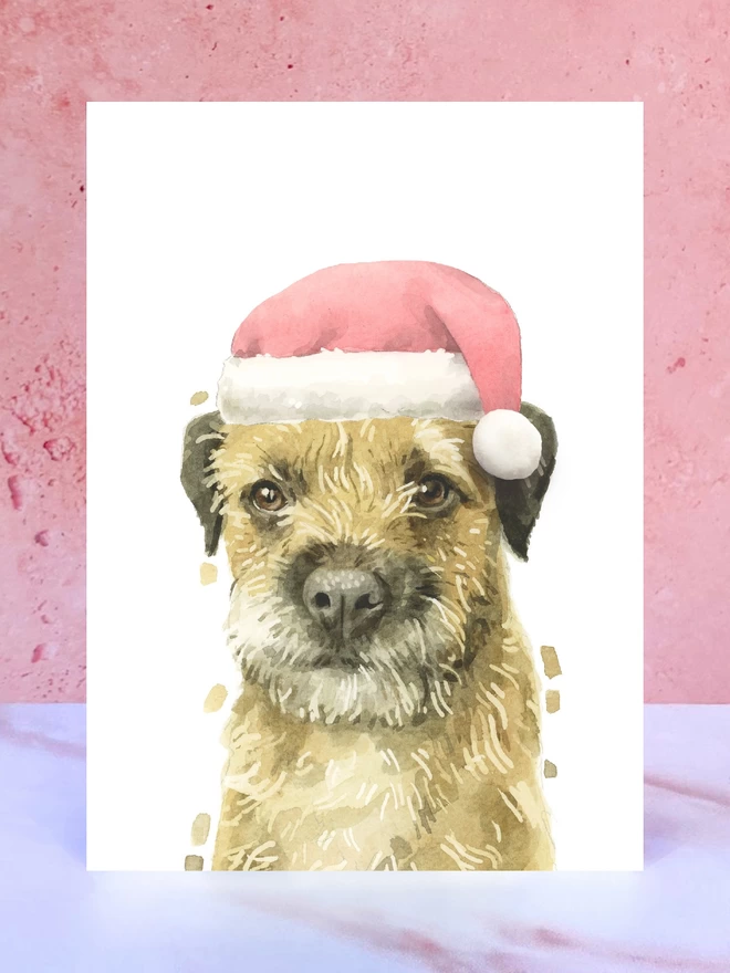 A Christmas card featuring a hand painted design of a border terrier, stood upright on a marble surface surrounded by pompoms. 