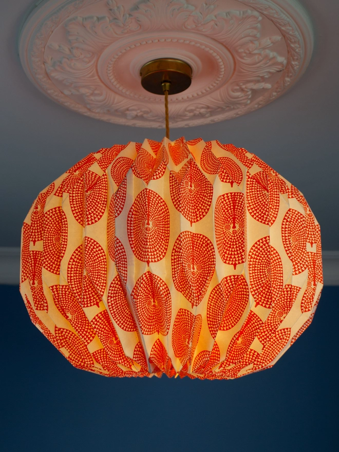 A vintage-inspired paper lampshade with a bold geometric pattern adds a touch of retro flair to a room with a blue wall and ornate ceiling. The lampshade is made from folded paper, creating a unique and eye-catching design.